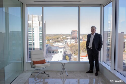Man with short grey hair wearing a dark grey suit and white dress shirt standing with hands in his pockets next to a white chair with an orange pillow in an office with large windows overlooking a city street and buildings.