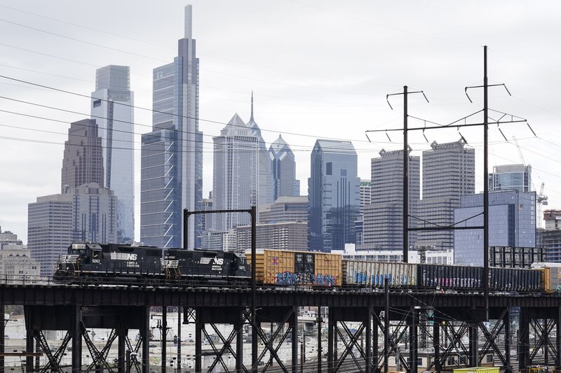 A freight train traverses elevated tracks with tall city buildings in the background