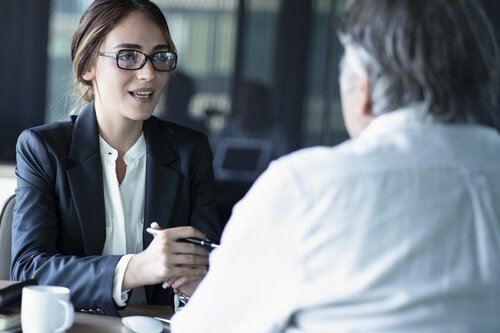 A young professional woman wearing glasses and a dark blazer sits at a desk and speaks with man, whose back is to the camera, in a modern office setting.