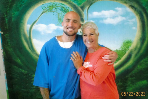 Bald man with a short beard wearing a loose blue shirt standing with his arm around a woman with short white hair wearing a coral long-sleeve top, both smiling in front of a painted mural of a heart-shaped tree opening to a blue sky background